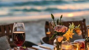 Een gedekte tafel met een glas rode wijn, kaarsen en bloemen, met uitzicht op een strand en golven.