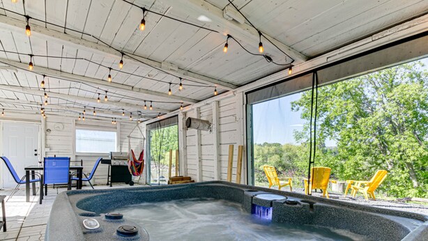 Un jacuzzi con revestimiento de piedra, rodeado de mobiliario exterior y amplias ventanas con vistas a árboles.