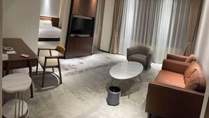 A hotel room with a bed, a TV, a dining table with chairs, a sofa, a coffee table, and a trash can.