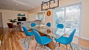 A modern living room with a glass dining table, blue chairs, a sofa, and a TV. - Emerald Dream, Oceanside House in Emerald Isle, NC (Emerald Isle)