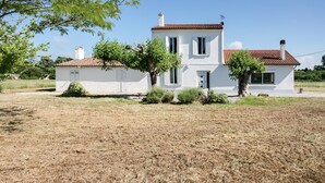 Une maison blanche à deux étages, surmontée d’un toit recouvert de tuiles rouges, entourée d’herbe sèche et d’arbres.
