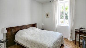 Une chambre à coucher avec une tête de lit en bois, un lit simple, une table de chevet avec une lampe, un petit tableau accroché au mur et une fenêtre avec des rideaux.