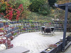 A paved patio with a metal table and chairs, surrounded by stone walls and various plants. - Vacation home near Bayernpark (Simbach)