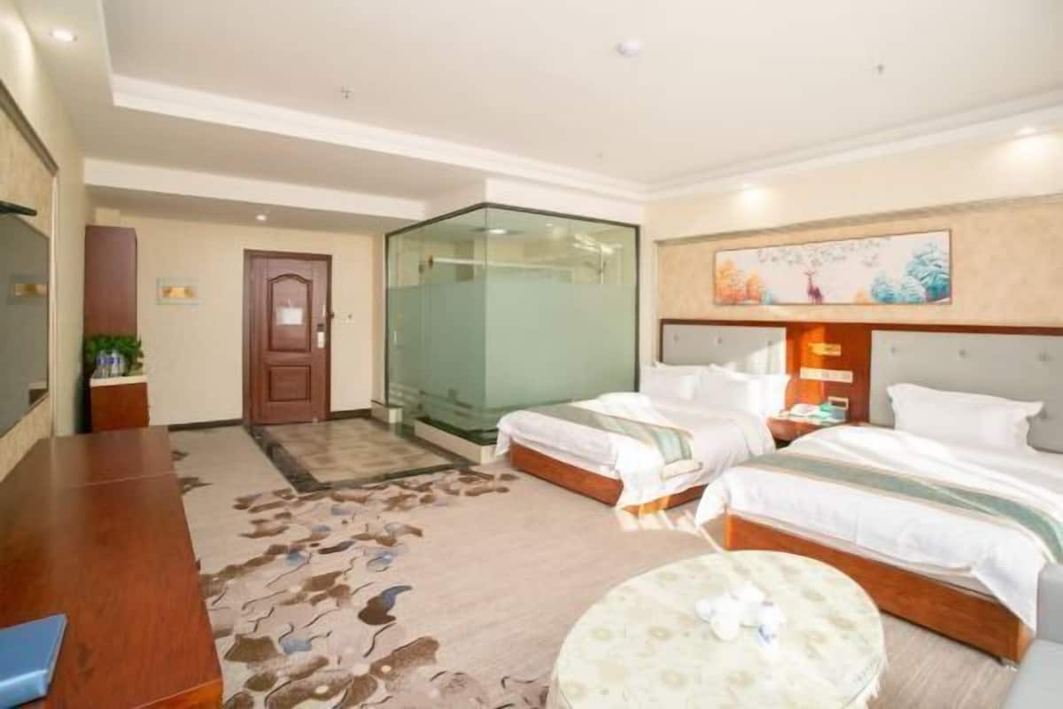 A hotel room with two beds, a desk, a chair, a small round table, a glass partition, and a painting on the wall.