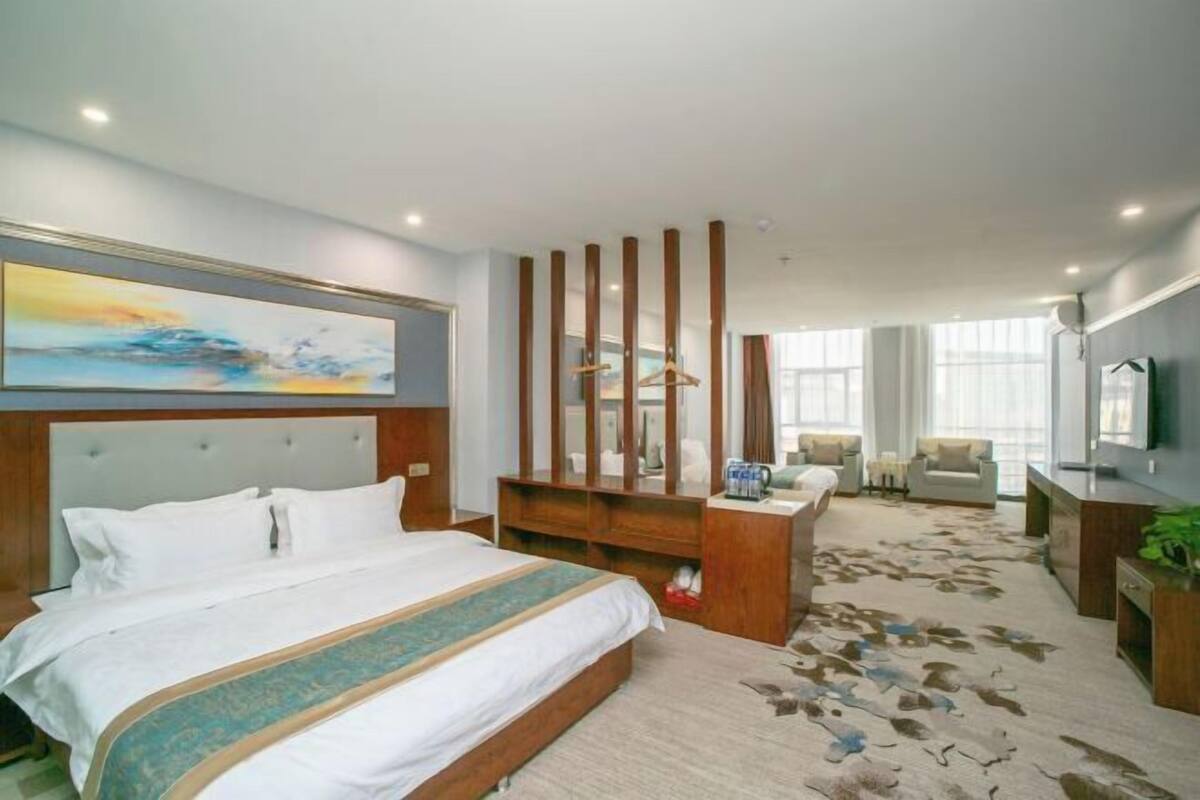 A hotel room with a large bed, a wooden headboard, a desk with a chair, a TV, and a large window.