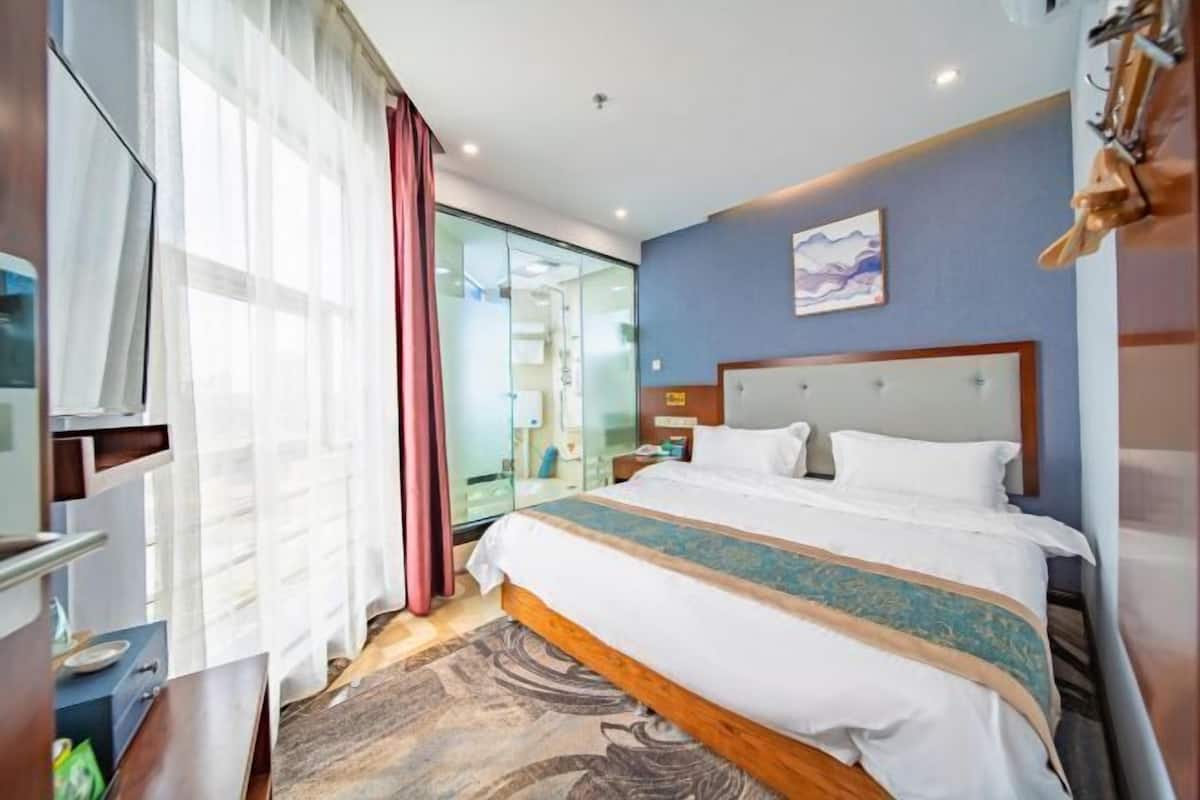 A hotel room with a bed, a TV, a desk, and a bathroom visible through a glass door.