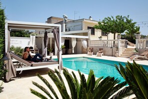 Zona de piscina con sillas de descanso, una pérgola y vista a un edificio al fondo.