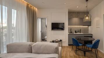 Una habitación de hotel moderna con cama, un escritorio pequeño, zona de comedor con sillas, televisor y área de cocina con lavamanos y encimera.