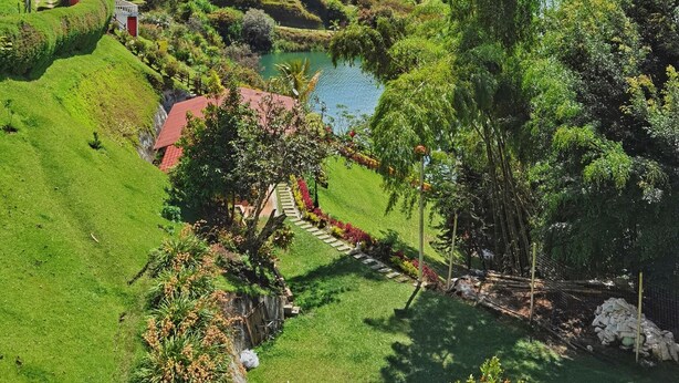 A lush green landscape with terraced fields, a small building with a red roof, and a body of water. - Descanso bajo la Piedra (Guatapé)