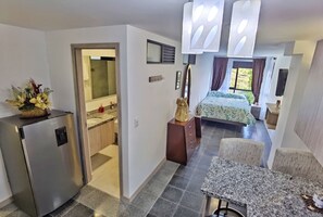 A compact hotel room with a kitchenette, a bed with a green patterned bedspread, a dining area with a granite table, and a large window with curtains.