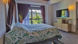 A bedroom with a bed, a TV, a window with curtains, and a view of greenery. - Descanso bajo la Piedra (Guatapé)