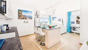 A modern kitchen with a central island, white bar stools, and a view of the sea through large windows.