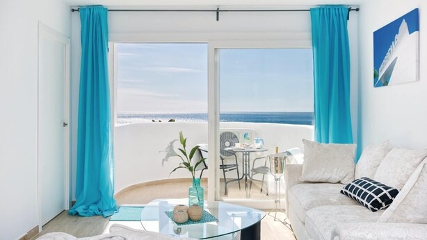 A room with a white sofa, a glass coffee table, and a view of the beach through a sliding glass door.