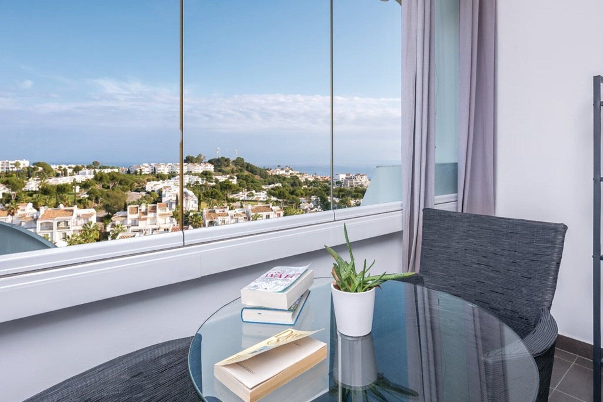 A balcony with a clear view of a coastal town, a glass table with books and a potted plant, and a dark chair.