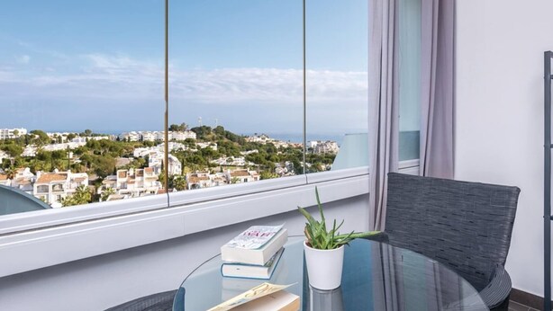 A balcony with a glass table, a wicker chair, a potted plant, and books. A cityscape is visible through the window. - Cosy apartment in Mijas (Mijas)