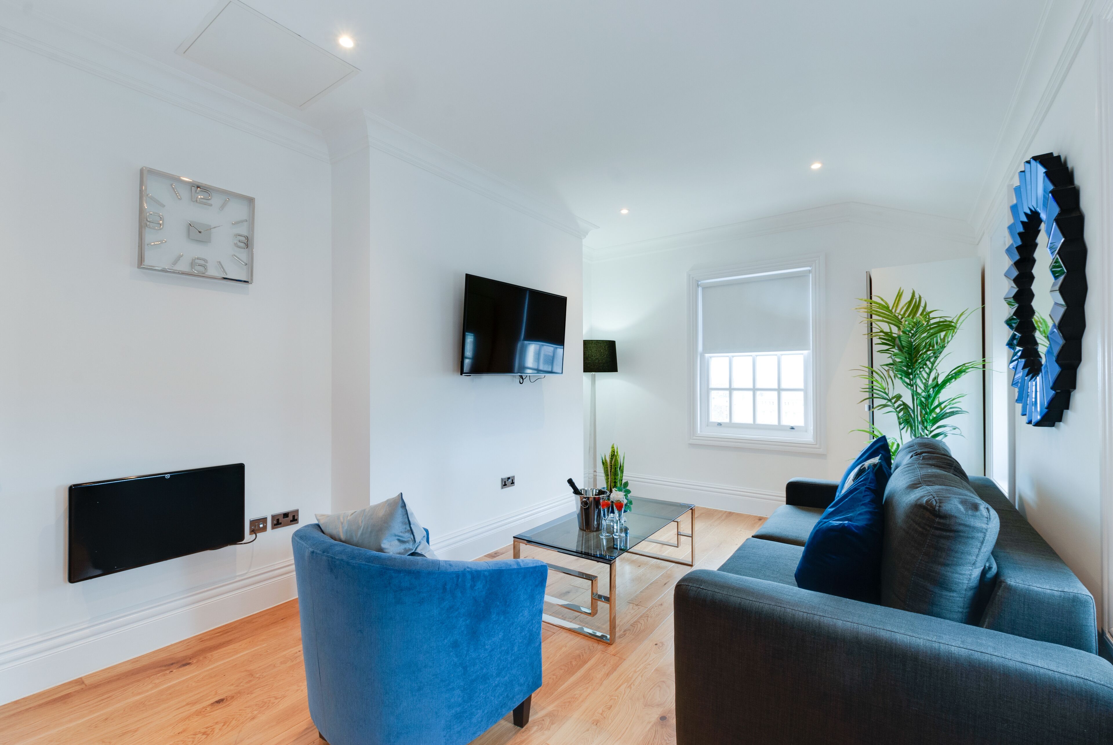 Una sala moderna con un sofá azul, un sofá gris, una mesa de centro de vidrio y un televisor de pantalla plana montado en la pared.