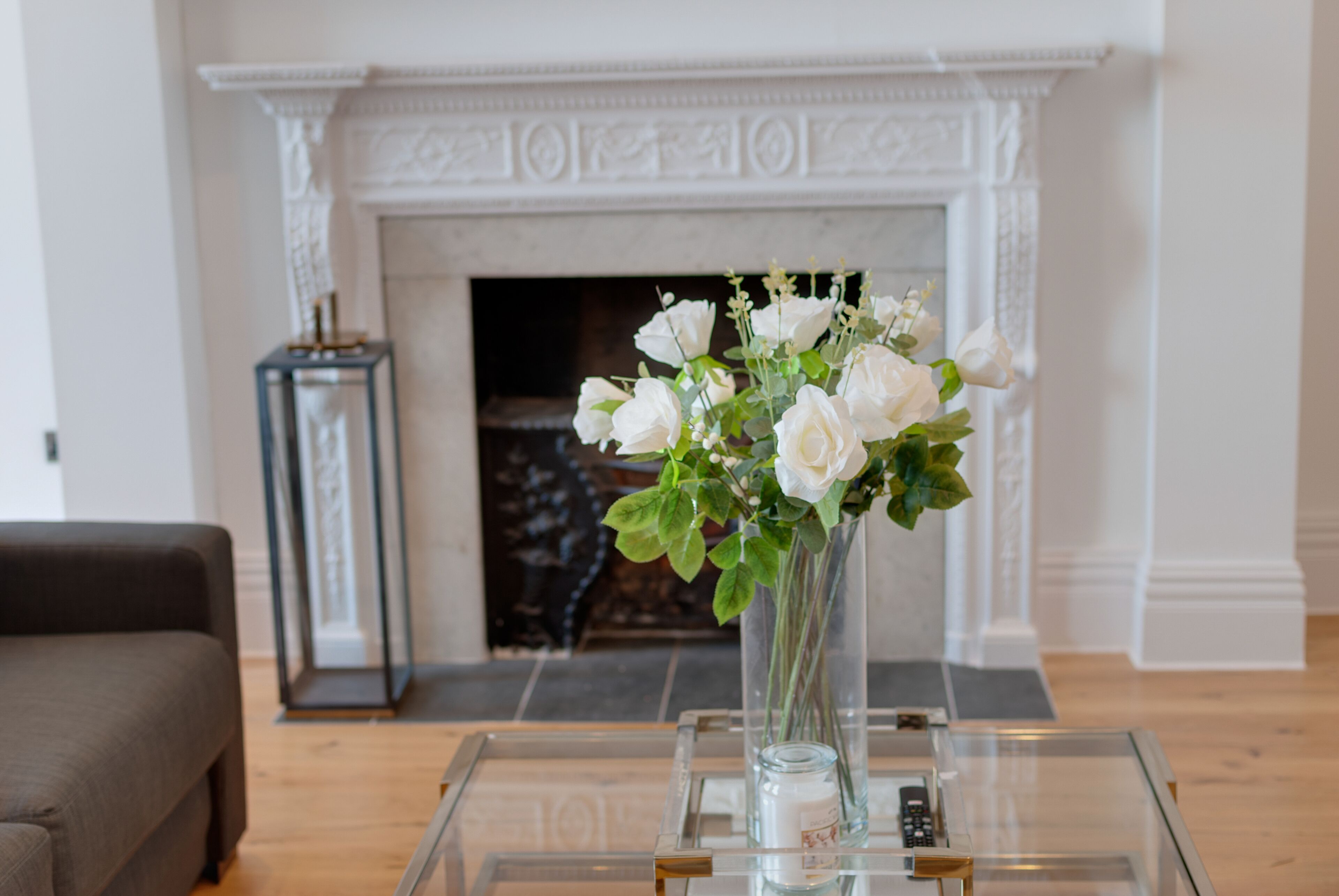 Un salon avec une cheminée, un vase de fleurs blanches posé sur une table en verre et un canapé marron.