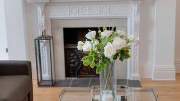 Un salon avec une cheminée, un vase de fleurs blanches posé sur une table en verre et un canapé marron.
