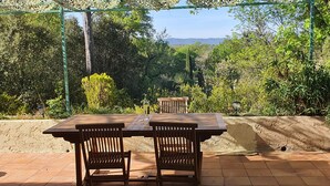 An outdoor patio with a wooden table and chairs, surrounded by greenery and a view of hills. - The Golden Gecko Country House  (Cotignac)