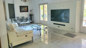 A modern living room with a white sofa, a glass coffee table, a dining area with chairs, a TV, and a chandelier. - The Golden Gecko Country House  (Cotignac)