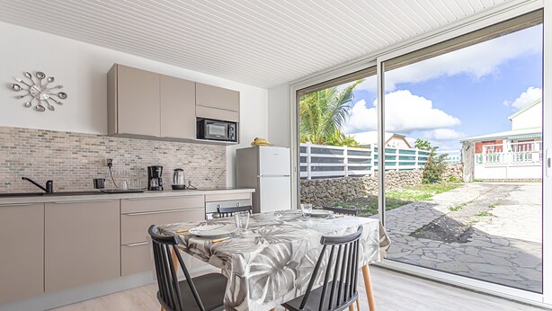 A modern kitchen with a dining area, featuring a large window, a white refrigerator, and a dining table with chairs. - Apartment La Désirade - Sainte-Anne (Sainte-Anne)