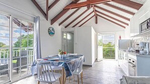 A spacious room with a dining table, chairs, and a kitchen area. There is a balcony with a view of greenery. - Les Saintes in Sainte-Anne (Sainte-Anne)