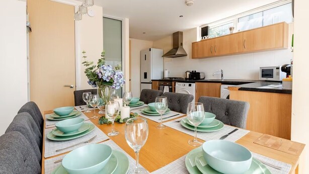 A modern dining area with a wooden table set for four, surrounded by grey sofas. A kitchen with wooden cabinets and a microwave is visible in the background. - Comfortable 3-Storey House with Free Car Parking (Manchester)