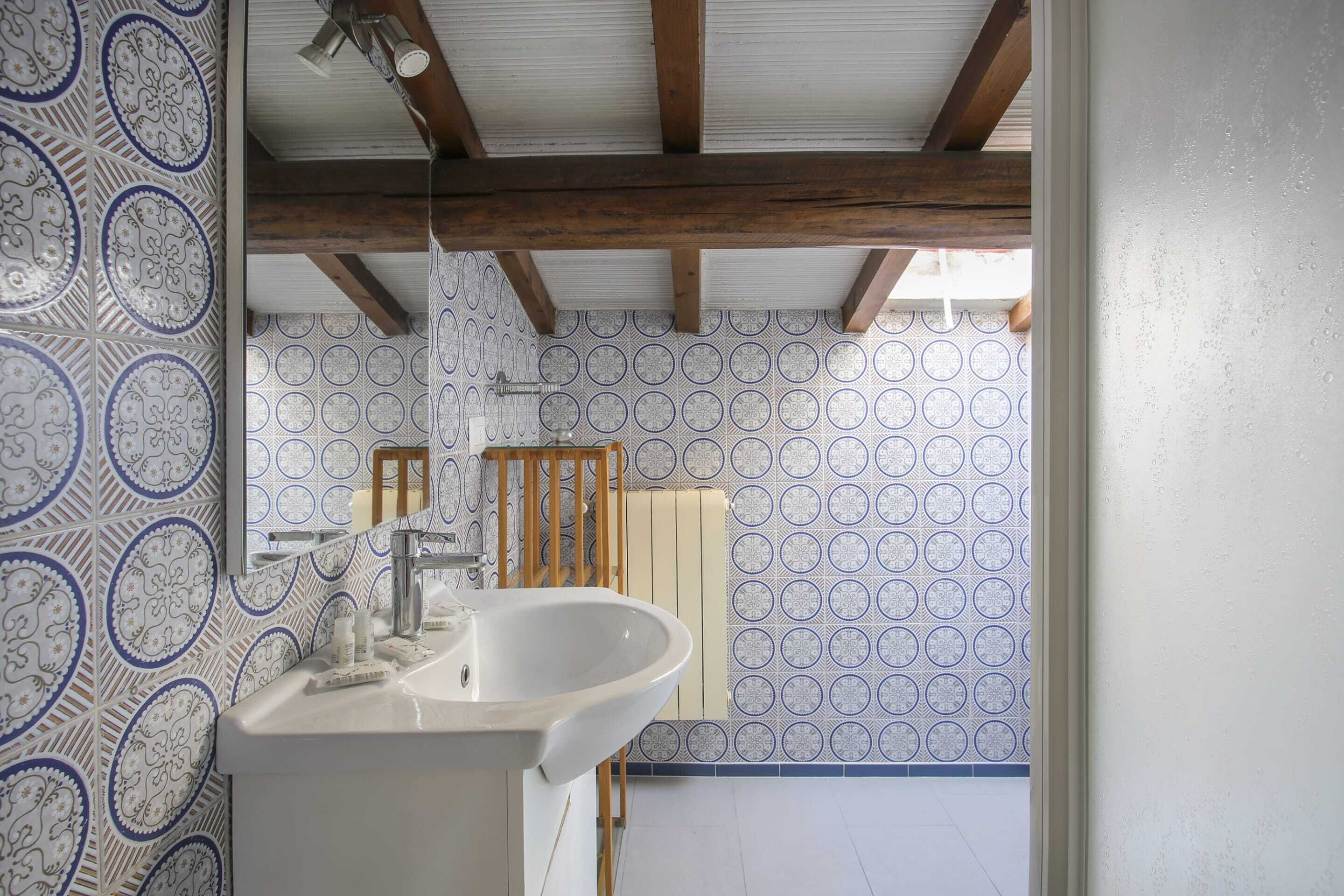 A bathroom with patterned tiles, a white sink, wooden beams, and a view of the outside.