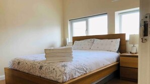 A neatly made bed with a patterned bedspread and pillows, a wooden headboard, a nightstand with a lamp, and a window providing natural light. - VERY CLEAN + AIR CONDITIONING + PARKING + INTERNET (Manchester)