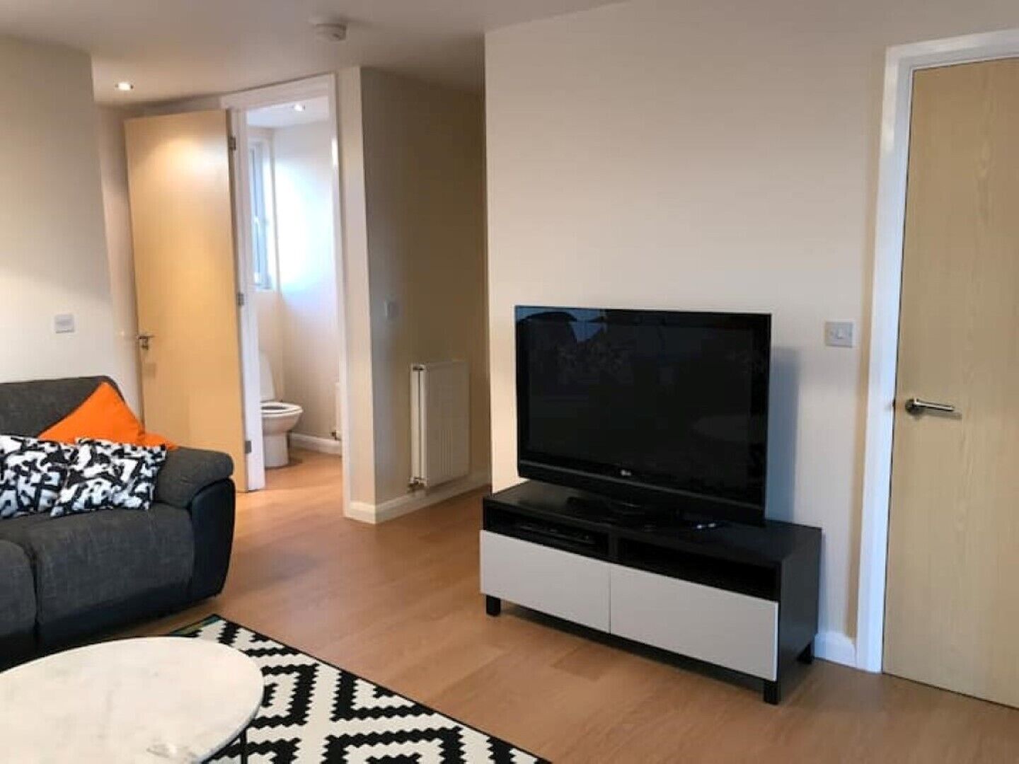 A modern living room with a flat-screen TV on a black stand, a grey sofa with an orange pillow, and a patterned rug.