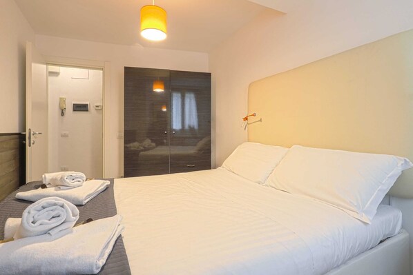 A hotel room with a bed, a bedside table with towels, a wardrobe, and a door with a phone and a light switch.