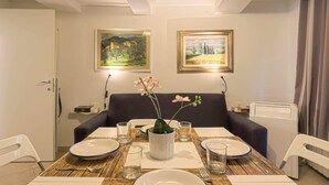 A dining area with a table set for two, a vase with flowers, and two framed paintings on the wall.