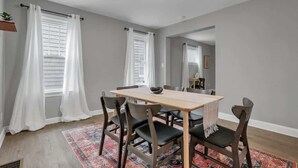 A dining area with a wooden table and chairs, a patterned rug, and white curtains on windows.