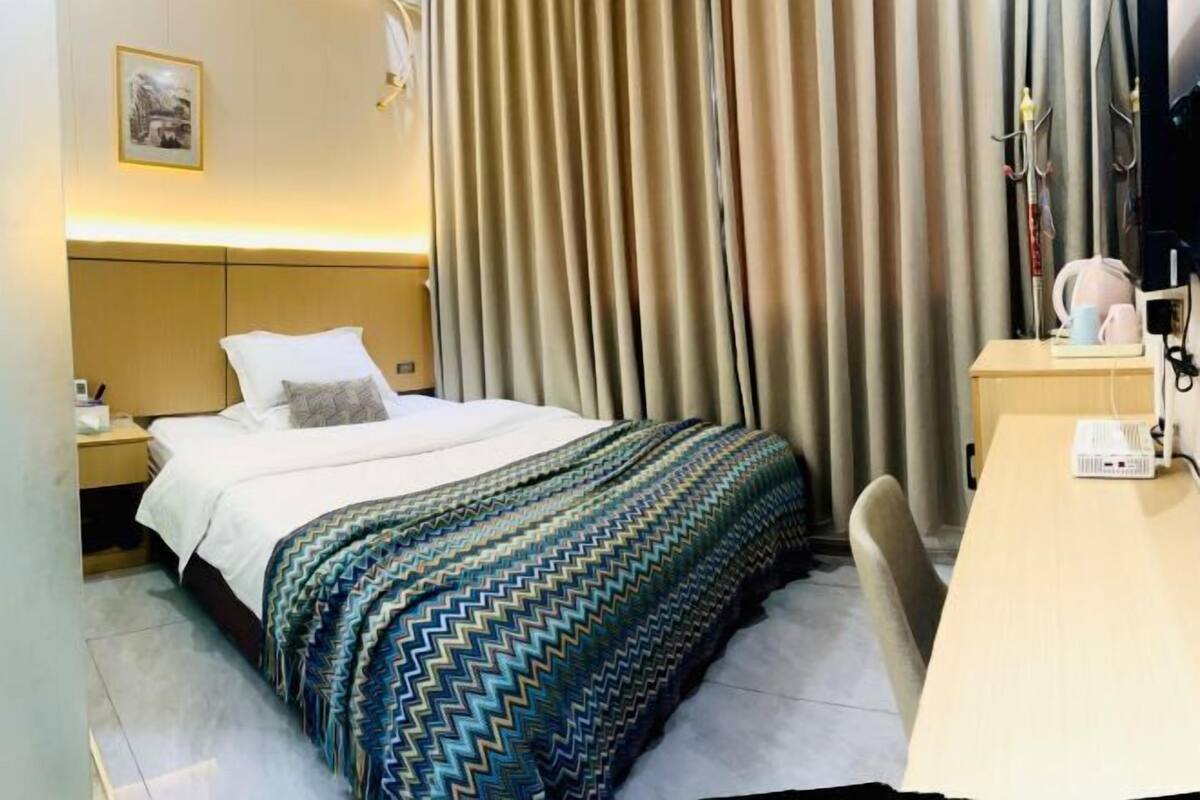Habitación de hotel con cama, escritorio, silla, cortinas, mesita de noche, lámpara y televisor instalado en la pared.
