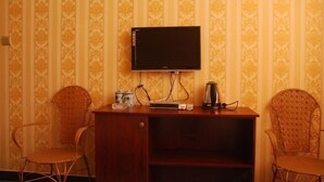 Una habitación con un televisor montado en la pared, un escritorio de madera con una tetera y dos sillones de mimbre.