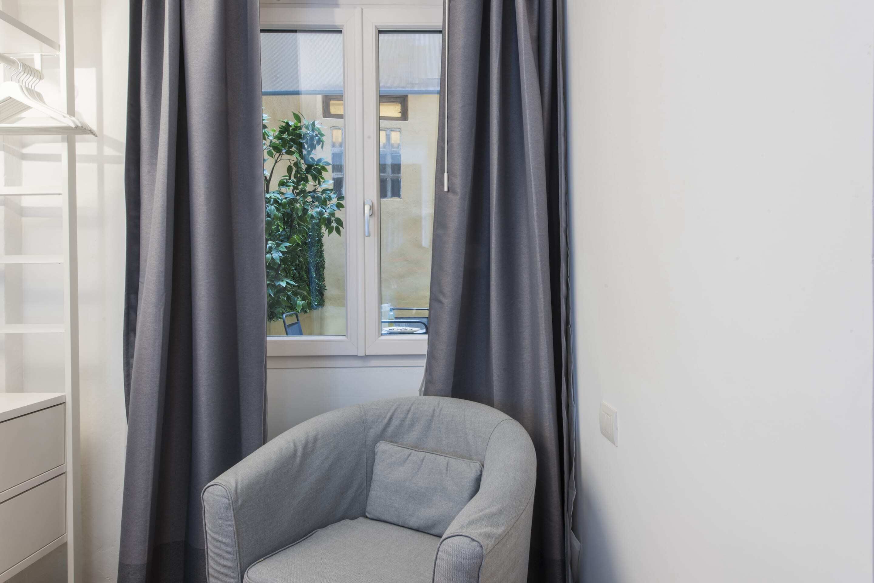 A corner room with a grey armchair, a window with curtains, and a view of a building outside.