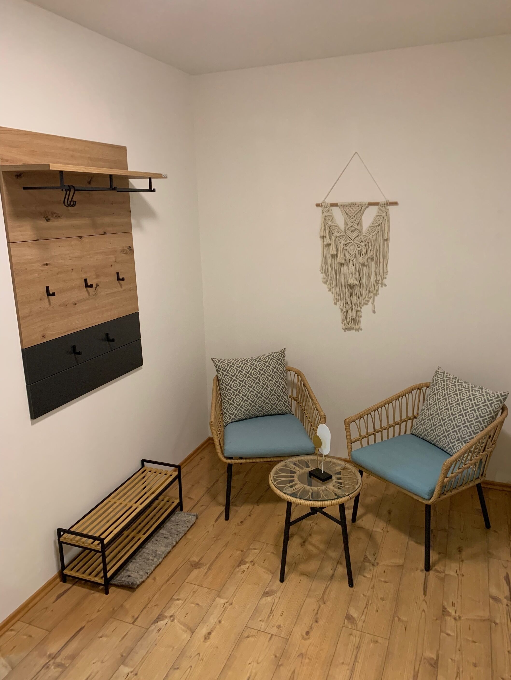 Een kamer met een houten bankje, twee rieten stoelen met gestreepte kussens, een klein rond tafeltje, een macramé wanddecoratie en een houten kapstok met haken.