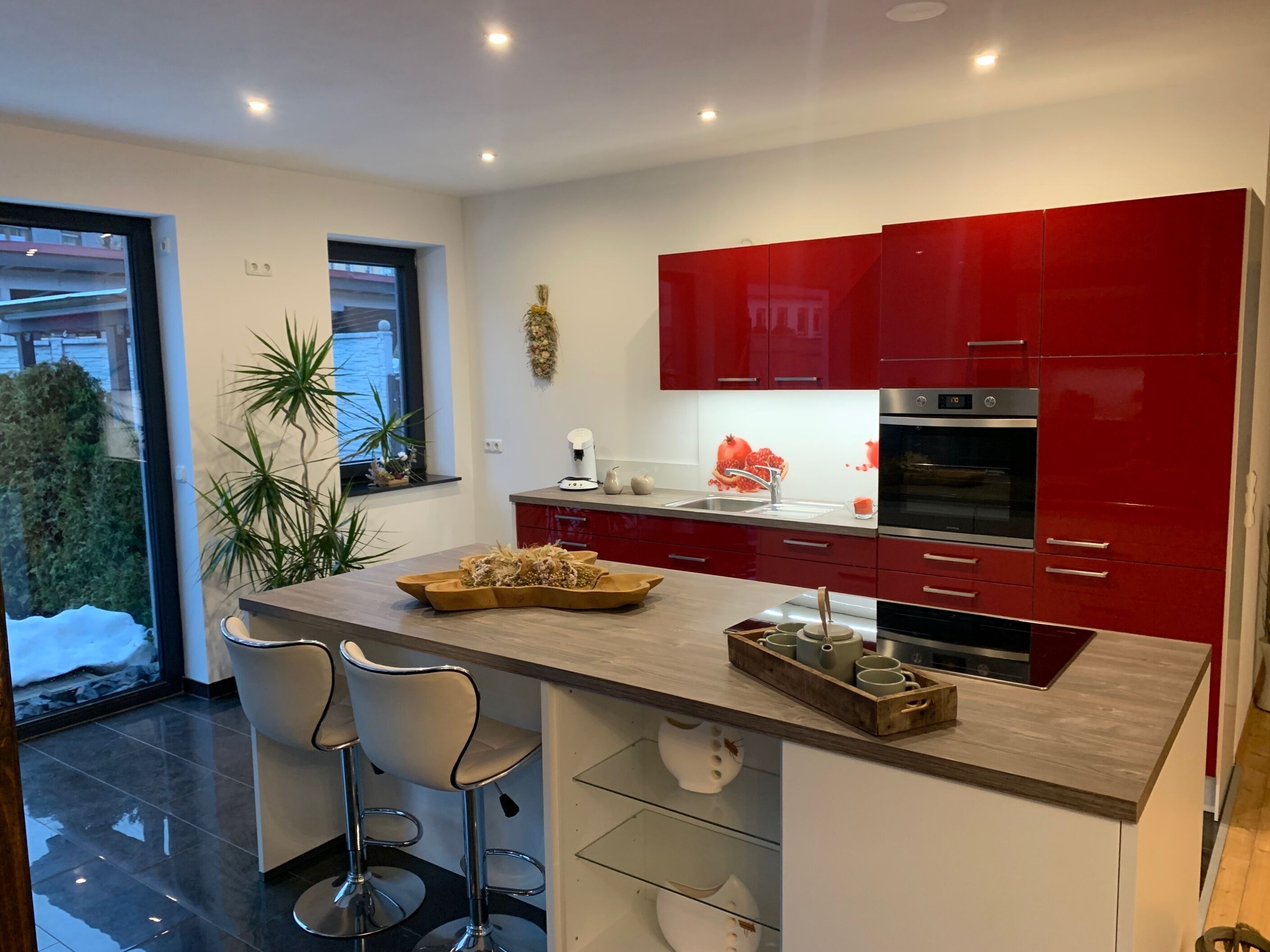 A modern kitchen with red cabinets, a central island with bar stools, and a view of the outdoors.