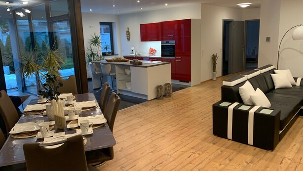 A modern living room with a dining area, a kitchen with red cabinets, and a black leather sofa. - "Körperzeit 1" Vacation Rental, Accessible (Lauscha)