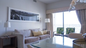 Une chambre d’hôtel moderne avec une table en verre, un canapé, deux fauteuils, un grand tableau encadré au mur et une vue sur l’extérieur par la fenêtre.