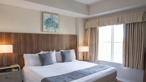 Une chambre d’hôtel avec un grand lit, deux lampes de chevet, une tête de lit en bois, une fenêtre avec des rideaux et une œuvre d’art florale accrochée au mur.