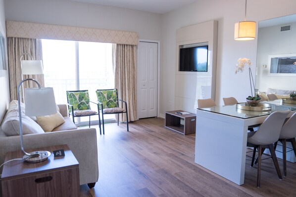 A modern hotel room with a sofa, a dining area with a glass table, a TV mounted on the wall, and a view of the outdoors through large windows. - 503 Luxury 1Bedroom pool and Universal view (Orlando)
