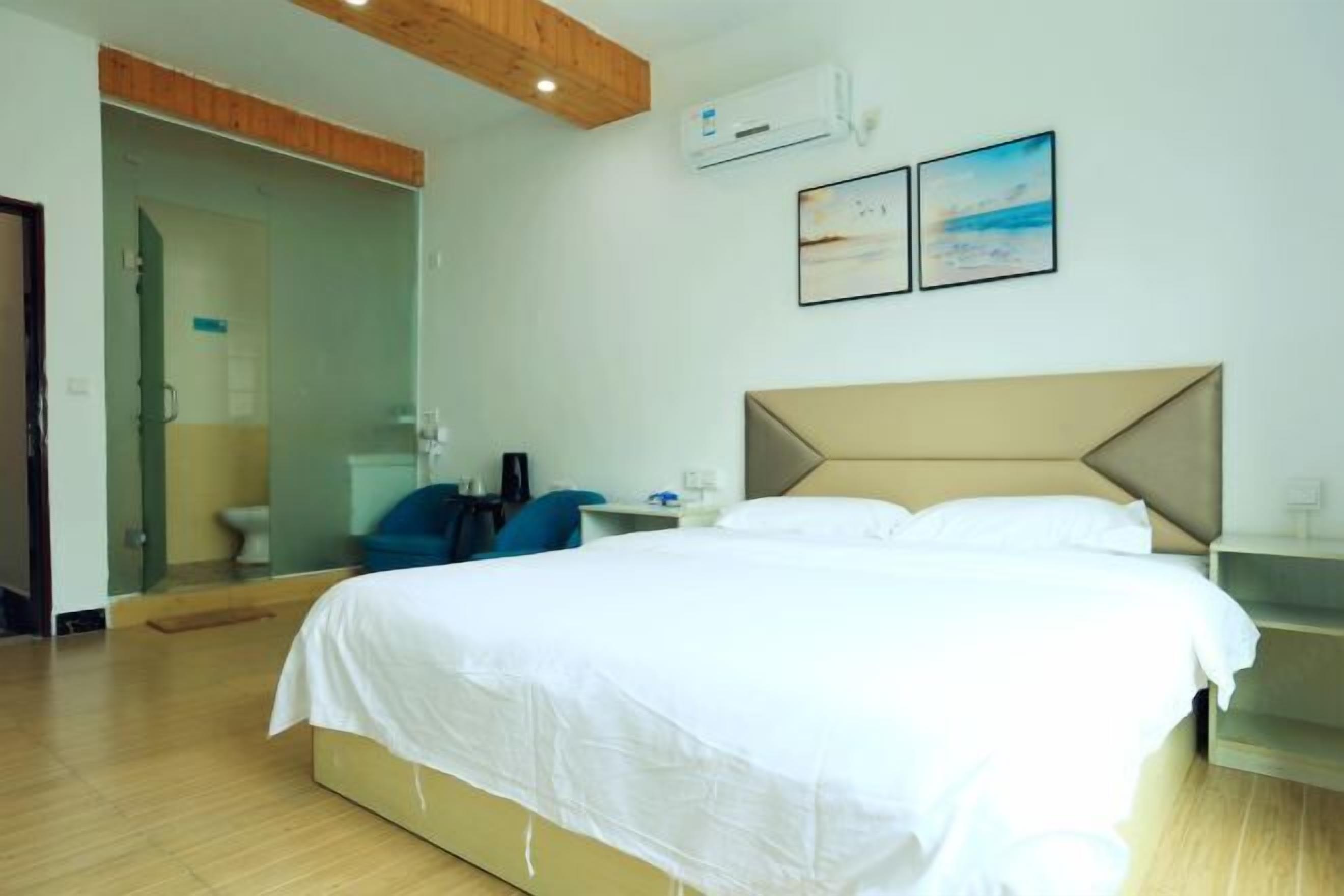 A hotel room with a bed, a desk, a chair, an air conditioning unit, and a glass door leading to a bathroom.