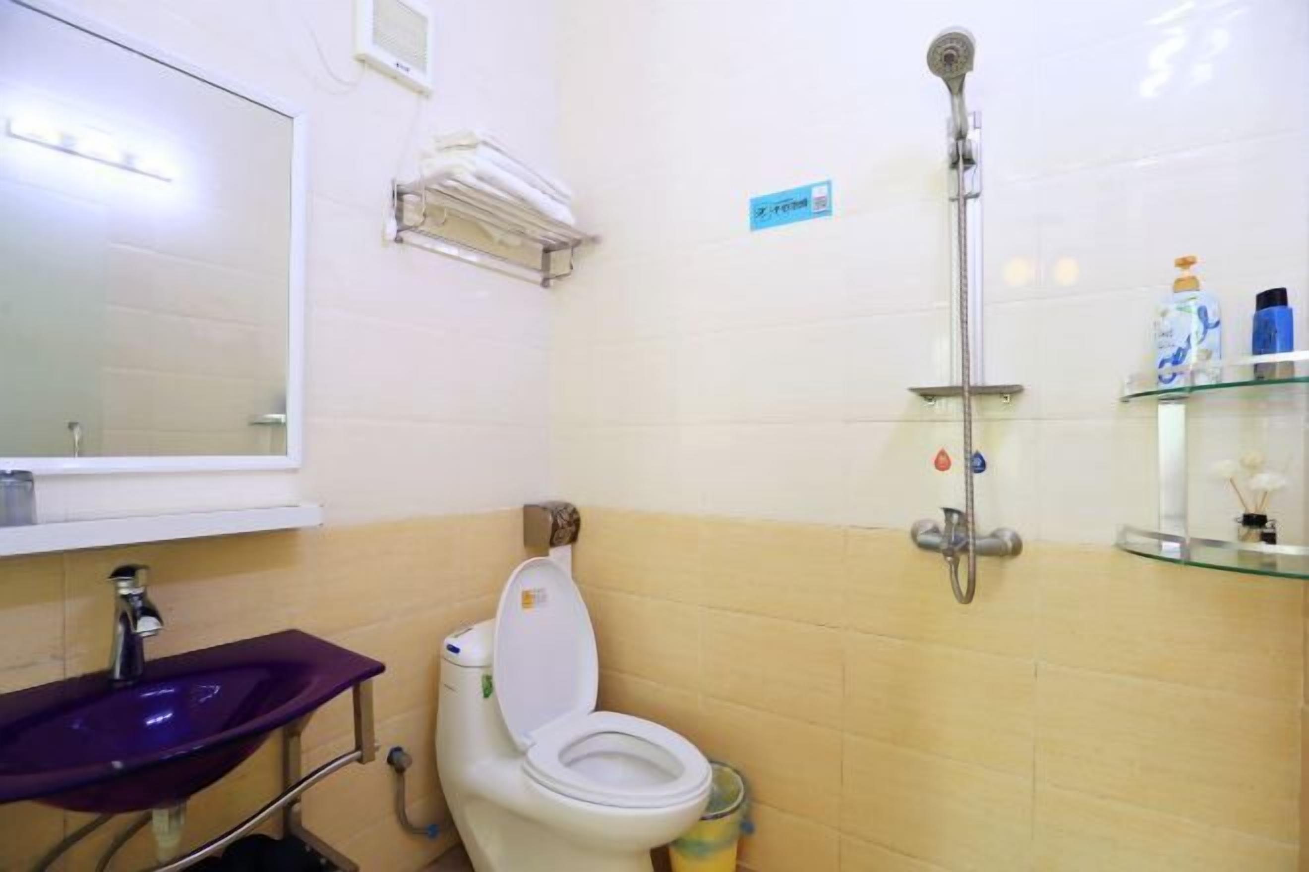 Un baño con un lavamanos morado, inodoro blanco, cabina de ducha de vidrio y un estante pequeño con artículos de aseo.