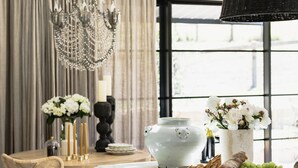 Une pièce bien éclairée, dotée d’une grande fenêtre, d’un lustre et d’une table sur laquelle se trouvent divers objets, notamment un vase de fleurs, des bougies et un pot décoratif.