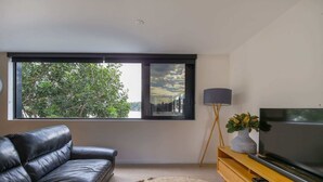 En moderne stue med en lædersofa, et fladskærms-tv på et træstel, et stort vindue med udsigt over byen og en gulvlampe.