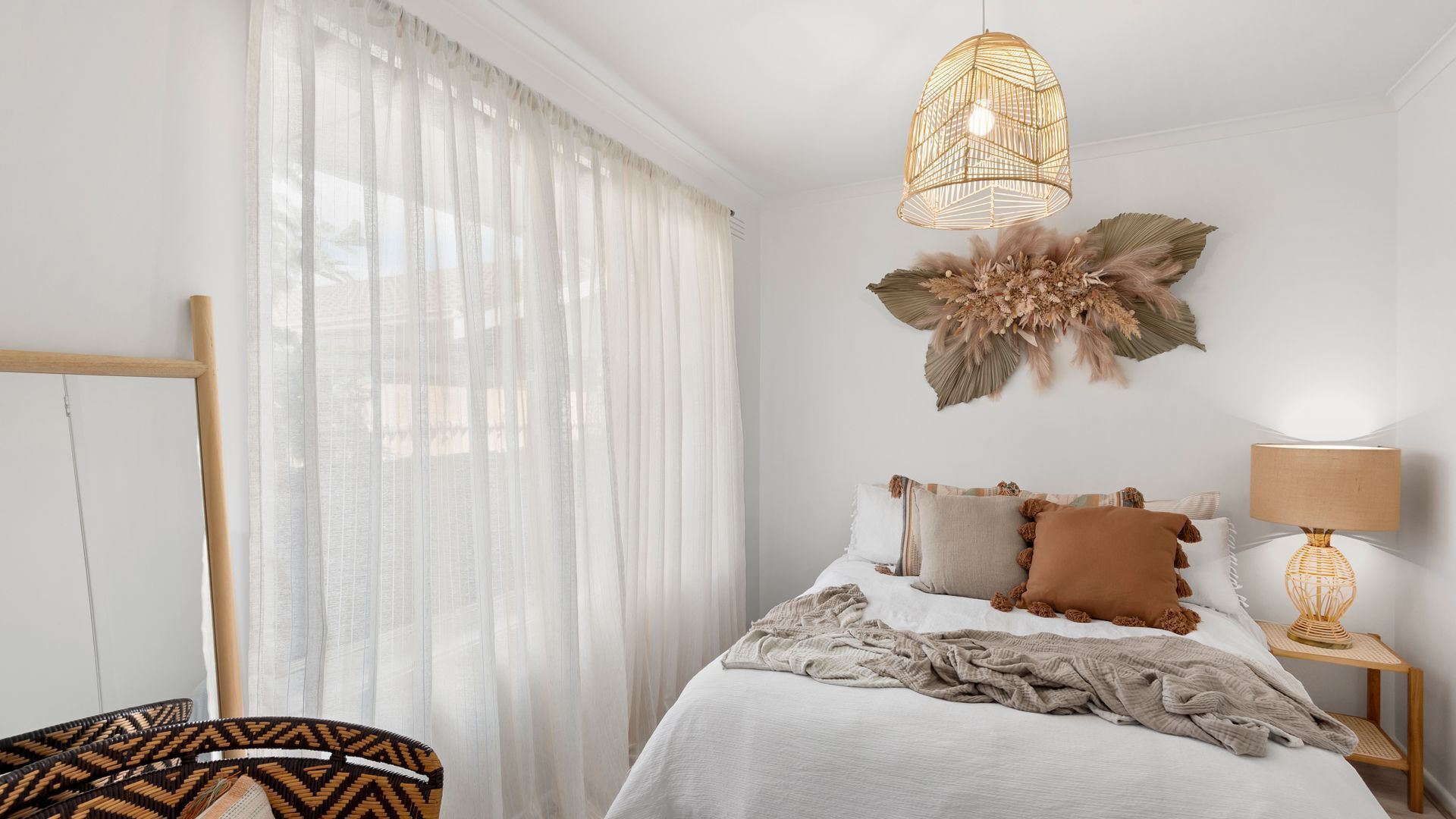 Ein Schlafzimmer mit einem Bett, einem Nachttisch mit Lampe, einem Sessel und einem großen Fenster mit durchsichtigen Vorhängen.