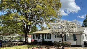 A white house with green shutters, a brick driveway, and a well-maintained garden with a large tree and blooming flowers. - Peaceful Place in Winston Salem (Winston-Salem)