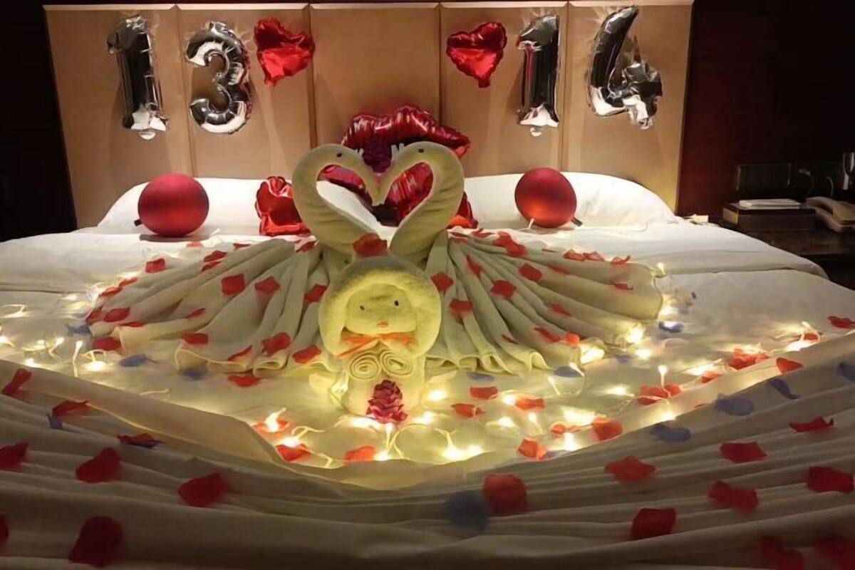 Una cama con un centro decorado con forma de cisne, rodeada de pétalos de rosa y luces, con globos con forma de corazón y los números '13' y '14' al fondo.