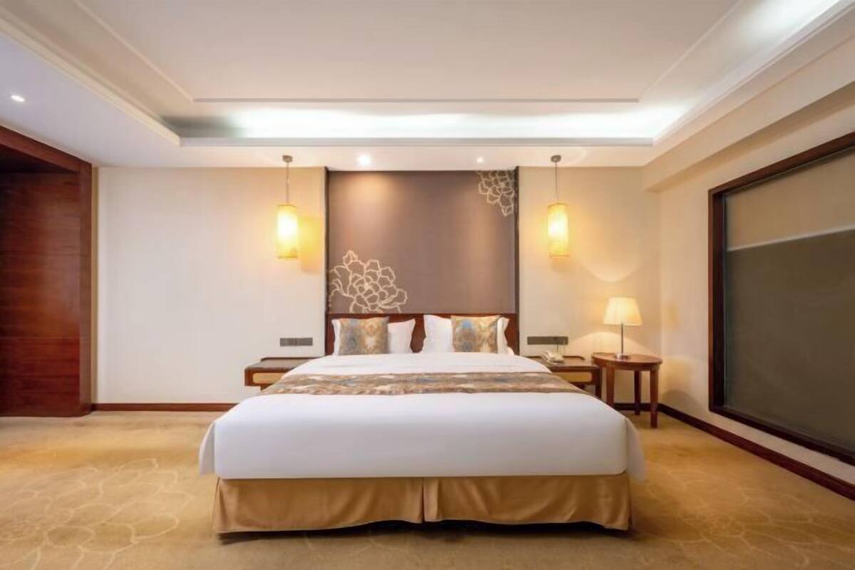 Habitación de hotel con una cama grande, dos mesitas de noche, una lámpara de pared y un ventanal amplio.